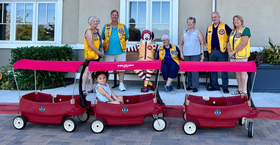 Seven Lions members in gold vests standing behind three red wagons outside Ronald McDonald House, with a child sitting in the middle wagon and a Ronald McDonald statue behind them.