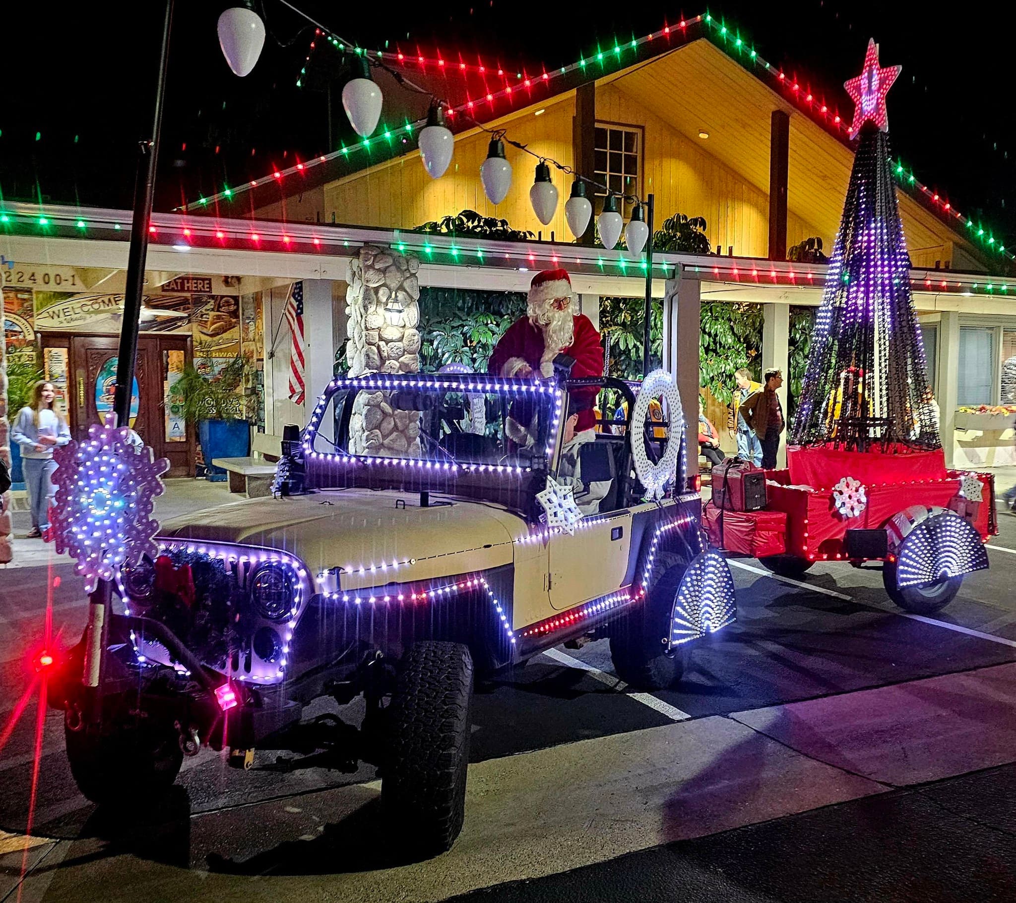 Santa riding in a Jeep strung with lights, towing a trailer of wrapped presents, outside Woody's Classic Grill at night.